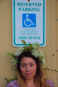 Emma, founder of Patient Perspective, poses in front of a handicap parking sign. She is wearing a crown made of flowers and smiles softly for the camera.